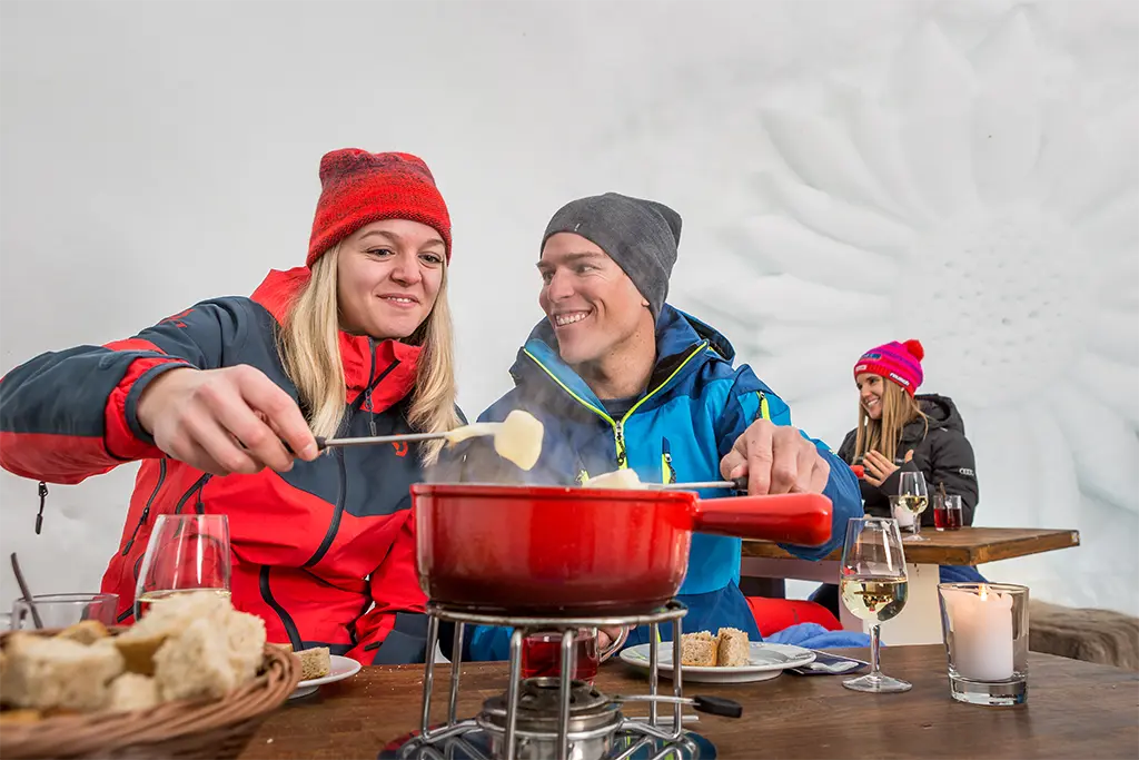 Mehrere Teilnehmer in einem großen Iglu beim Fondue-Essen im Rahmen einer Fondue Party als Teil eines Winter Teamevents in der Schweiz. Die Teilnehmer haben Spaß und genießen das Winterincentive