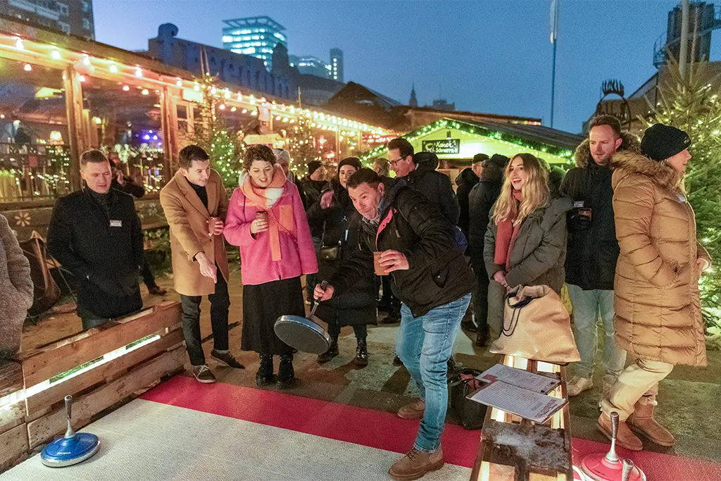 Eine Eventgruppe beim Eisstockschießen als Teil eines Firmen Weihnachtsmarkt auf Outdoorbasis. Ein Teilnehmer ist mit einem Curling aktiv und die anderen Teilnehmer der Weihnachtsfeier feuern ihn bei dem Motivationsevent an