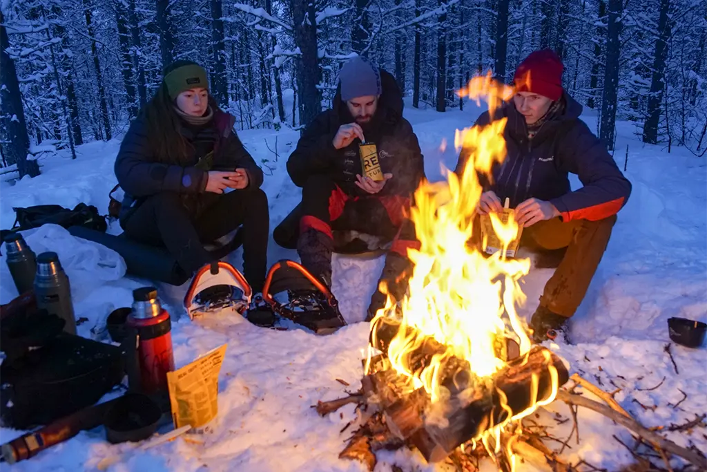 Drei Teilnehmer eines Outdoorevents im Schnee rund um eine Feuerstelle als Teil eines Winterteamevents mit Outdooraktivitäten im Schnee.
