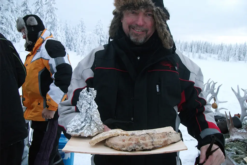 Ein Teilnehmer bei einem Winterevent mit einem Lachs auf einem Brett als Teil eines Outdoor-Teamkochevents im Winter. Der Teilnehmer lacht und hat Spaß an dem Firmenevent im Schnee.