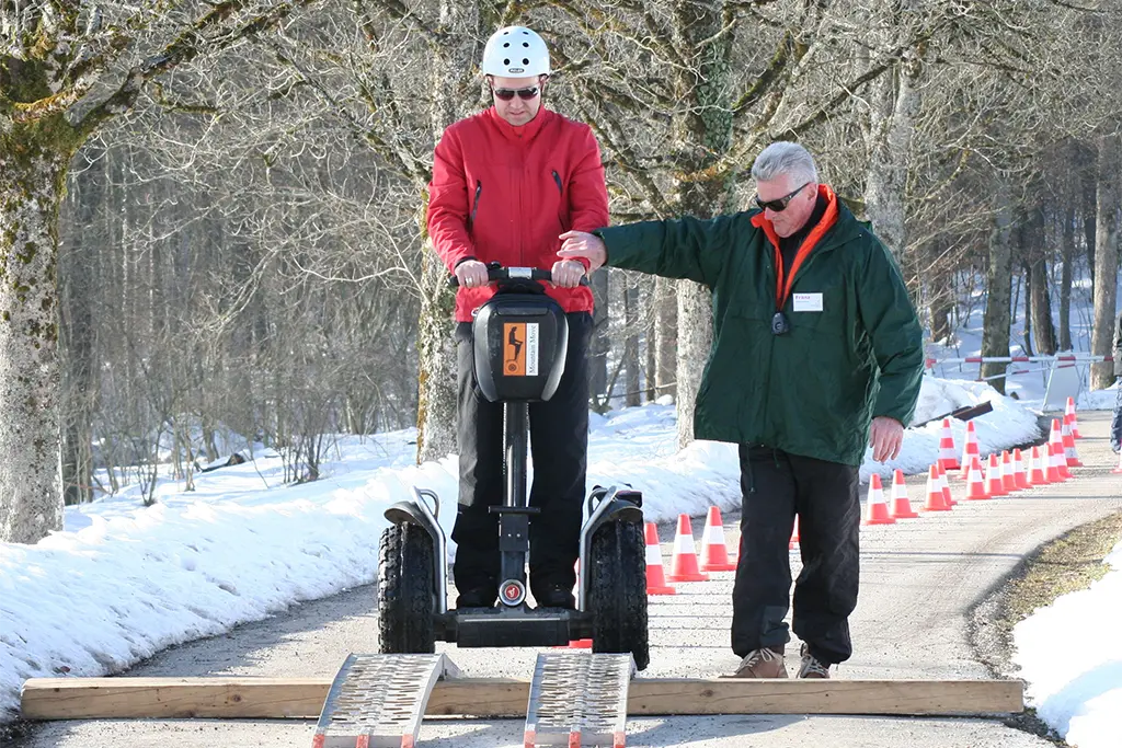 Ein Teilnehmer fährt unter Beobachtung eines Instruktors des Segway-Veranstalter team projekt bei einem Winter Teamparcours mit einem Segway über ein Hindernis.