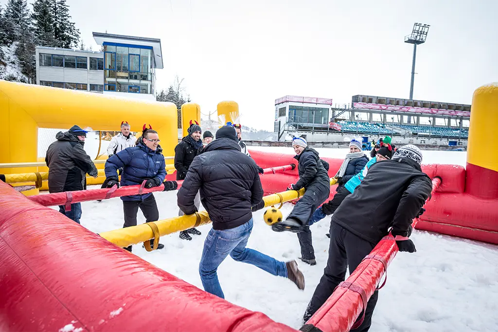 Teilnehmer spielen in einer Human-Table-Anlage Fußball im Schnee und sind begeistert. Das Fußballspiel im Schnee ist Teil einer Teamolympiade im Winter im Allgäu.