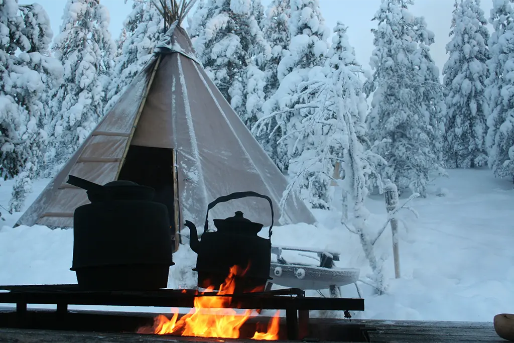 Ein Tipi im Schnee und einer Feuerstelle mit Glühwein-Töpfen als Teil eines Winter-Teamevents im Schnee. Im Hintergrund sieht man den verschneiten Schwarzwald und Teilnehmer, die Spaß an dem Teambuilding Programm haben.