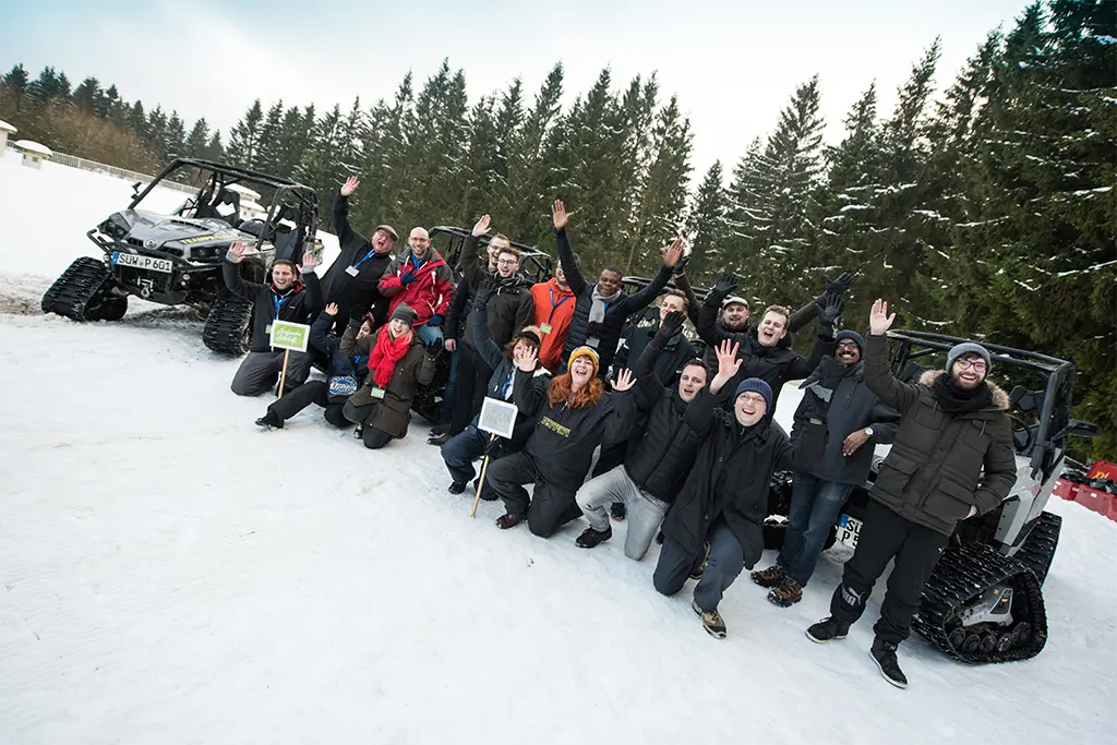 Eine jubelnde Gruppe im Schnee mit Schneemobilen bei einer Winter-Teamolympiade am Tegernsee. Das Schneeevent wurde von der Eventagentur Die Erlebniscompany organisiert
