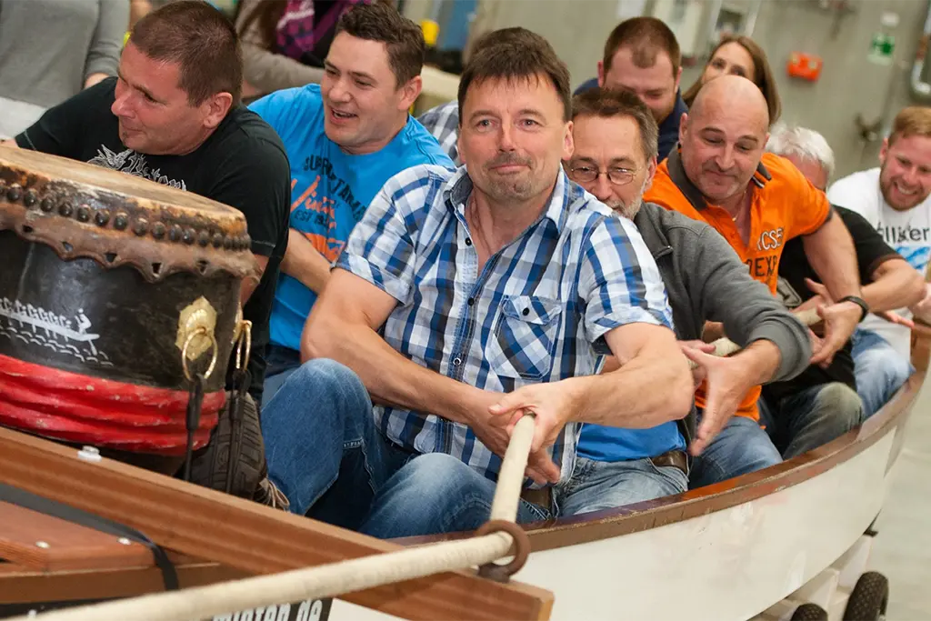Ein Indoor-Drachenbootrennen in einer Eventhalle in Frankfurt mit Teilnehmer, die in einem Drachenboot sitzen und bei einem Firmenevent Spaß haben. Die Teilnehmer des Indoorevents ziehen die Drachenboote am Tau zum Ziel